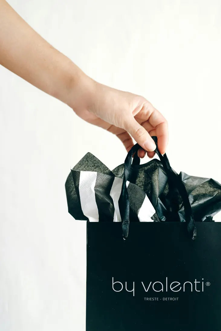A hand holding a black shopping bag with the By Valenti brand logo.