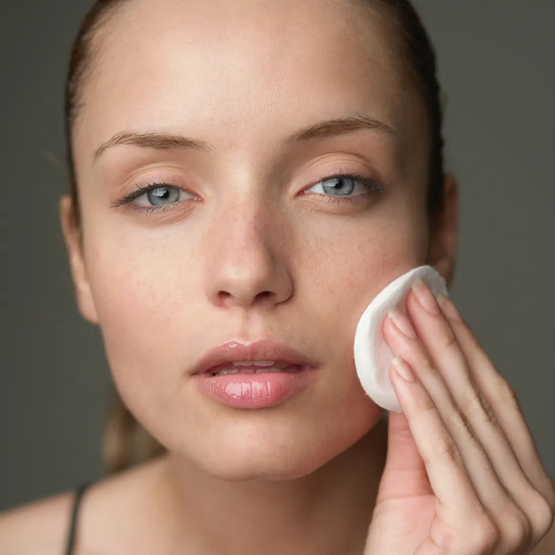 Woman applying a cosmetic product with a cotton pad to her face against a neutral background