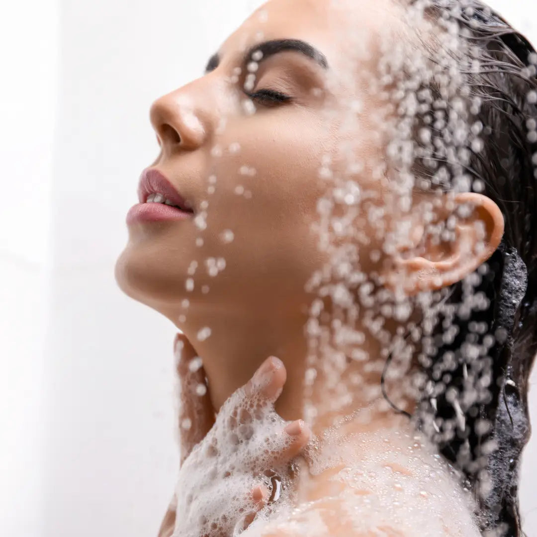 Woman showering with BY VALENTI bar soap. Suds on her face and shoulders, against a white background
